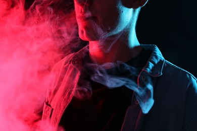 Photo of Man using electronic smoking device in neon lights on black background, closeup
