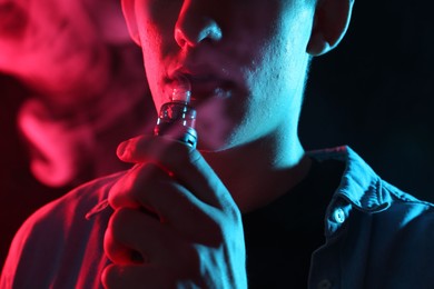 Photo of Man using electronic smoking device in neon lights on black background, closeup