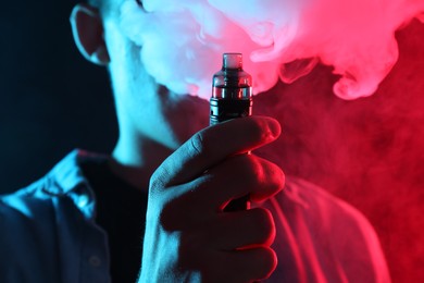 Photo of Man using electronic smoking device in neon lights on dark background, closeup