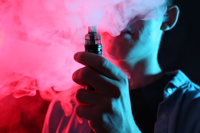 Photo of Man using electronic smoking device in neon lights on dark background, closeup