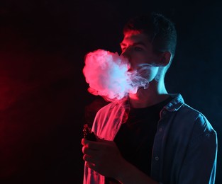 Photo of Man using electronic smoking device in neon lights on dark background, space for text