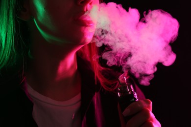 Photo of Woman using electronic smoking device in neon lights on black background, closeup