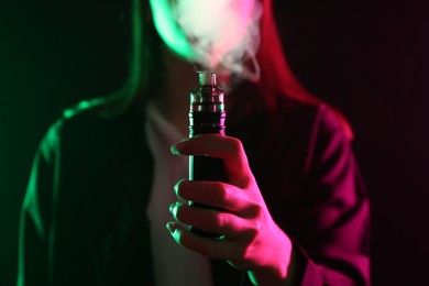 Photo of Woman using electronic smoking device in neon lights on black background, closeup