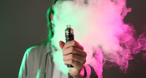 Photo of Woman using electronic smoking device in neon lights on grey background, selective focus