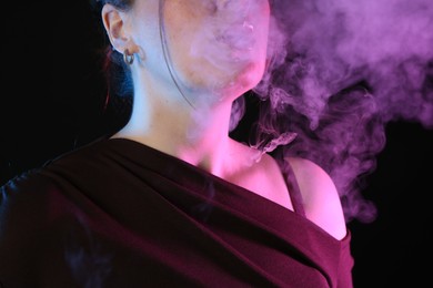 Photo of Woman using electronic smoking device in neon lights on black background, closeup