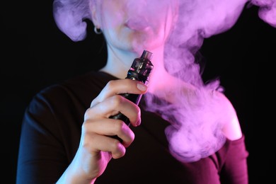 Photo of Woman using electronic smoking device in neon lights on black background, closeup