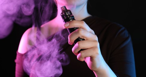 Photo of Woman using electronic smoking device in neon lights on black background, closeup