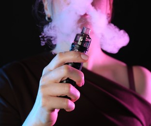 Photo of Woman using electronic smoking device in neon lights on black background, closeup