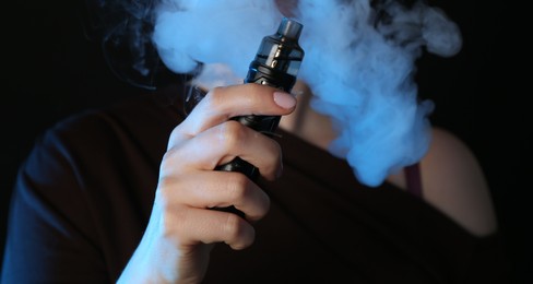 Photo of Woman using electronic smoking device in neon lights on black background, closeup