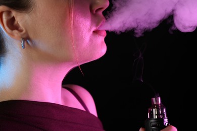 Photo of Woman using electronic smoking device in neon lights on black background, closeup