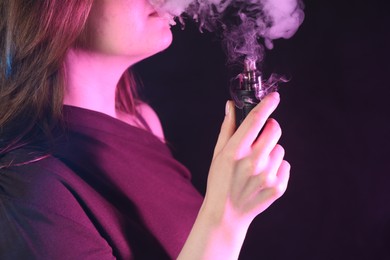 Photo of Woman using electronic smoking device in neon lights on black background, closeup