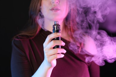 Photo of Woman using electronic smoking device in neon lights on black background, closeup