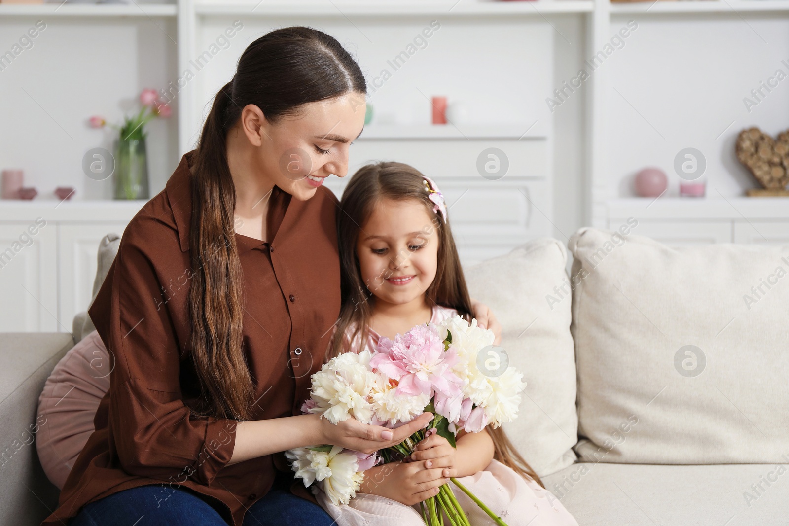 Mother and her daughter with bouquet of beautiful peonies on sofa at home Photo of Mother and her daughter with bouquet of beautiful peonies on sofa at home