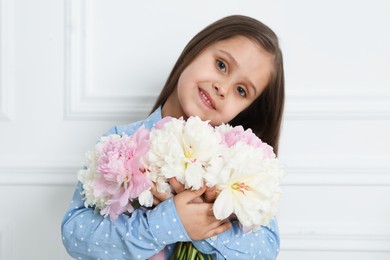 Photo of Cute little girl with bouquet of beautiful peonies near white wall indoors