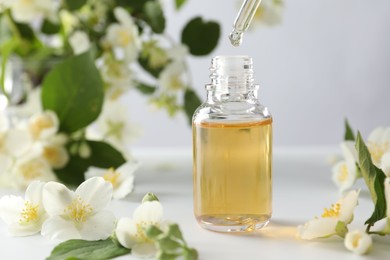 Photo of Dripping essential oil into bottle and beautiful jasmine flowers at white table, closeup