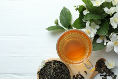 Photo of Aromatic jasmine tea in glass cup, flowers and leaves on white wooden table, flat lay. Space for text