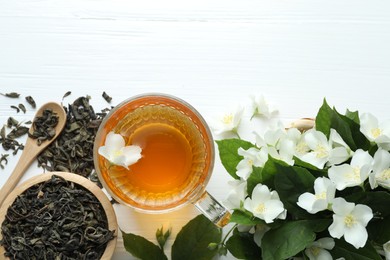 Photo of Aromatic jasmine tea in glass cup, flowers and leaves on white wooden table, flat lay