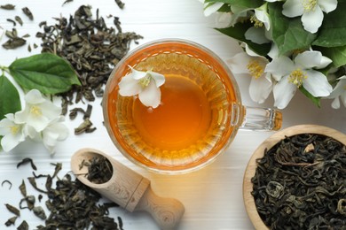 Photo of Aromatic jasmine tea in glass cup, flowers and leaves on white wooden table, flat lay