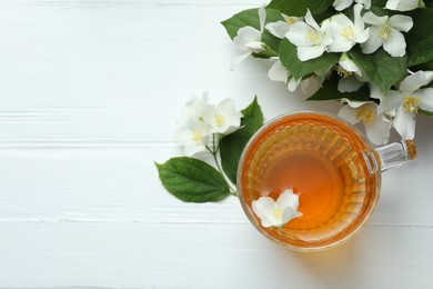Photo of Aromatic jasmine tea in glass cup, flowers and green leaves on white wooden table, flat lay. Space for text