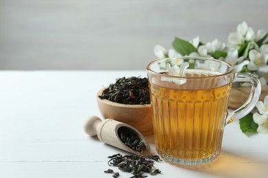 Photo of Aromatic jasmine tea in glass cup, flowers and dried leaves on white wooden table, closeup. Space for text