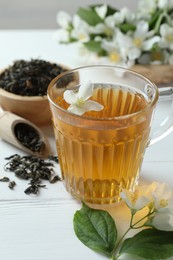 Photo of Aromatic jasmine tea in glass cup, flowers and leaves on white wooden table, closeup