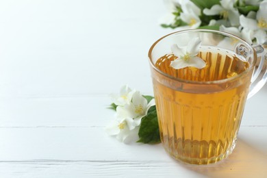 Photo of Aromatic jasmine tea in glass cup, flowers and green leaves on white wooden table, closeup. Space for text