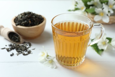 Photo of Aromatic jasmine tea in glass cup, flowers and dried leaves on white wooden table, closeup