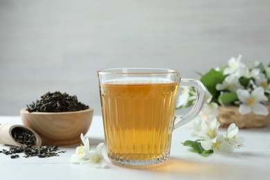 Photo of Aromatic jasmine tea in glass cup, flowers and dried leaves on white wooden table, closeup
