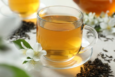 Photo of Aromatic jasmine tea in glass cup, flowers and dried leaves on white table, closeup
