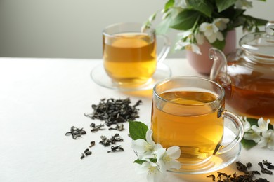Photo of Aromatic jasmine tea in glass cups, teapot, flowers and dried leaves on white table, closeup. Space for text