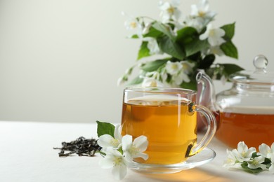 Photo of Aromatic jasmine tea in glass cup, teapot, flowers and dried leaves on white table, closeup. Space for text