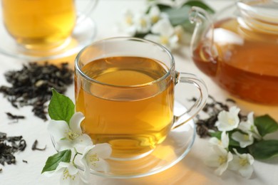 Photo of Aromatic jasmine tea in glass cup, teapot, flowers and dried leaves on white table, closeup