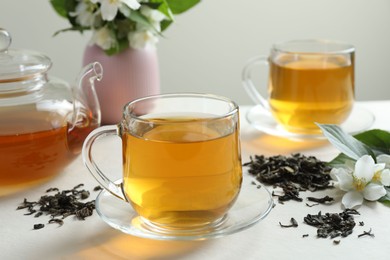 Photo of Aromatic jasmine tea in glass cups, teapot, flowers and dried leaves on white table, closeup
