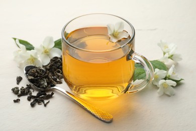 Photo of Aromatic jasmine tea in glass cup, flowers and leaves on white table, closeup