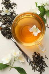 Photo of Aromatic jasmine tea in glass cup, flowers and leaves on white table, flat lay