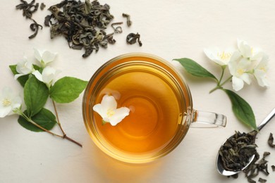 Photo of Aromatic jasmine tea in glass cup, flowers and leaves on white table, flat lay