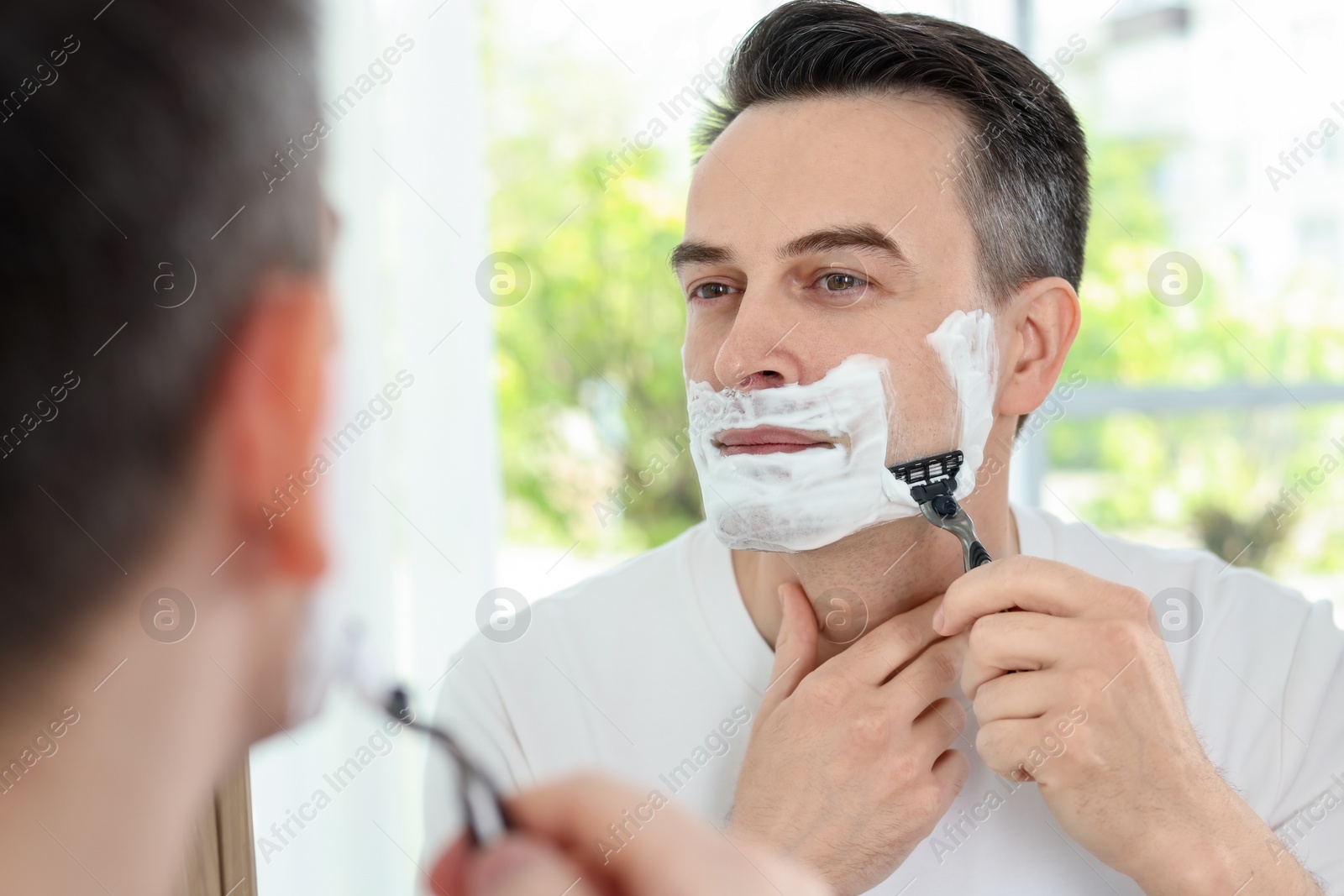 Handsome man shaving with razor near mirror at home Photo of Handsome man shaving with razor near mirror at home
