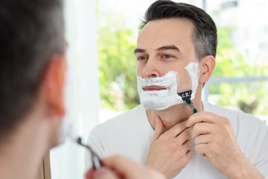 Photo of Handsome man shaving with razor near mirror at home