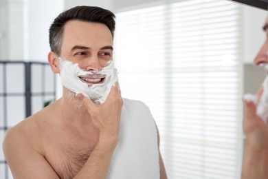Photo of Smiling man applying shaving foam near mirror in bathroom