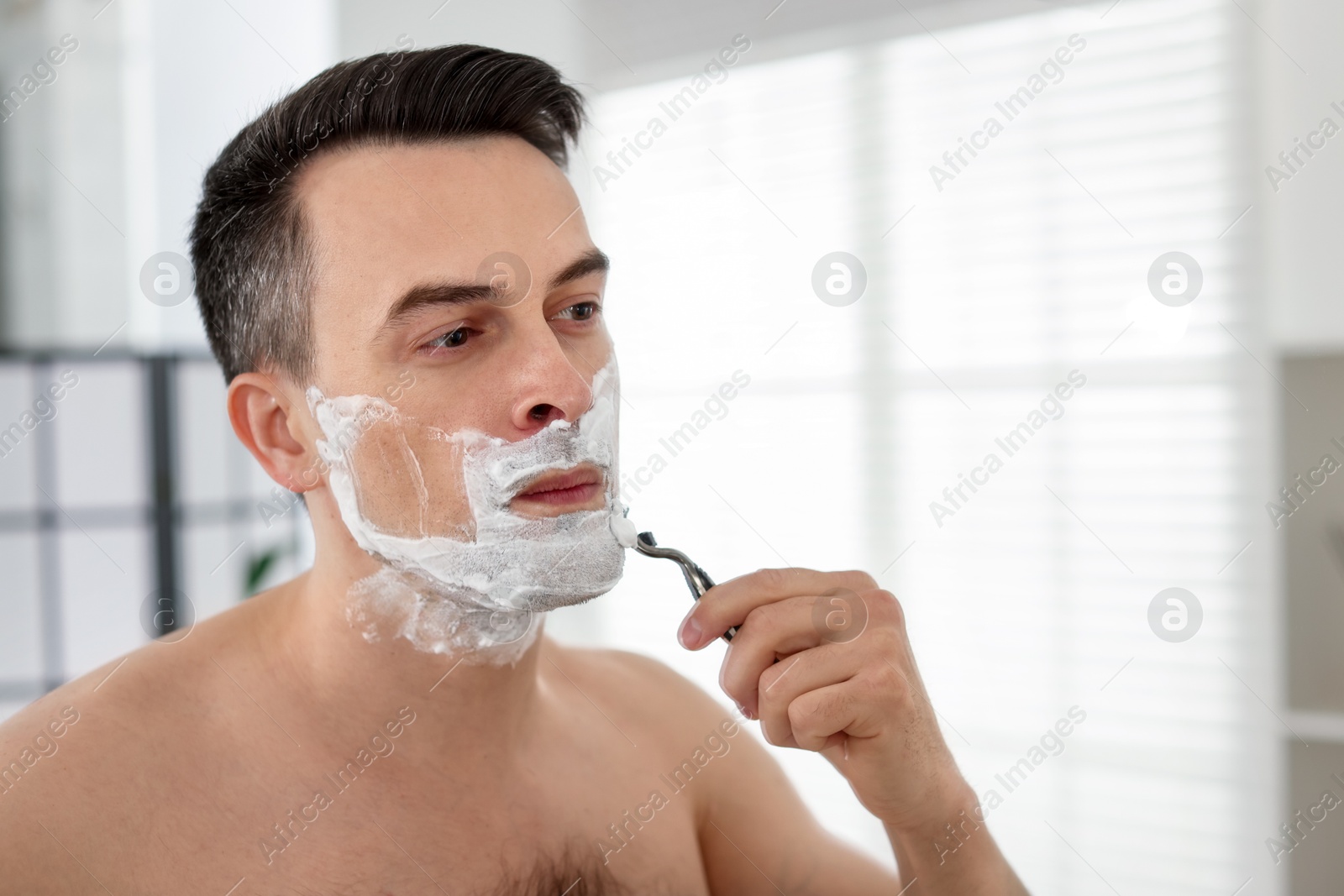 Handsome man shaving with razor near mirror at home Photo of Handsome man shaving with razor near mirror at home