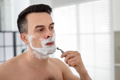 Photo of Handsome man shaving with razor near mirror at home