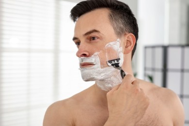 Photo of Handsome man shaving with razor near mirror at home