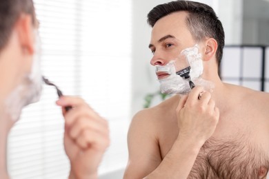 Photo of Handsome man shaving with razor near mirror at home