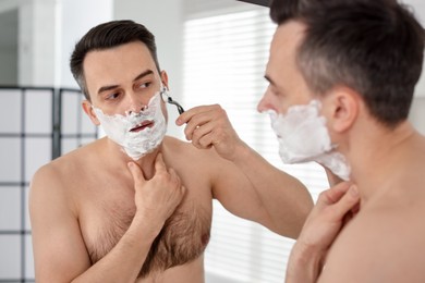 Photo of Handsome man shaving with razor near mirror at home