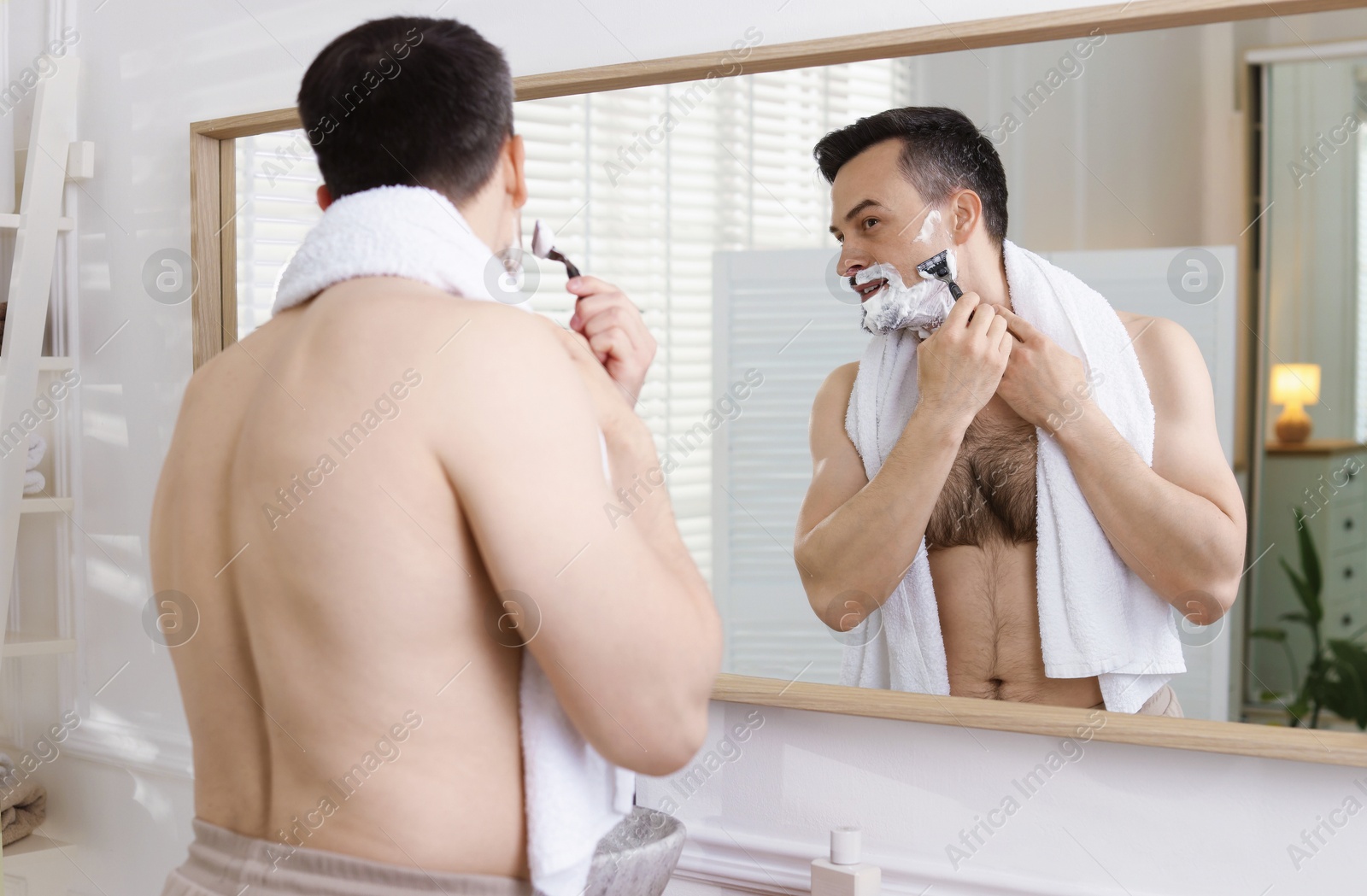 Handsome man shaving with razor near mirror at home Photo of Handsome man shaving with razor near mirror at home