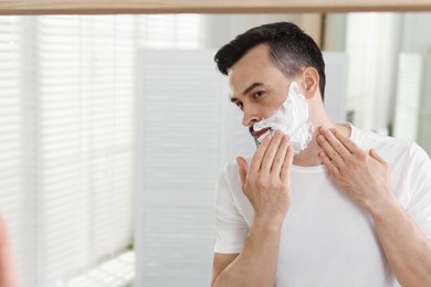 Photo of Handsome man applying shaving foam near mirror in bathroom. Space for text