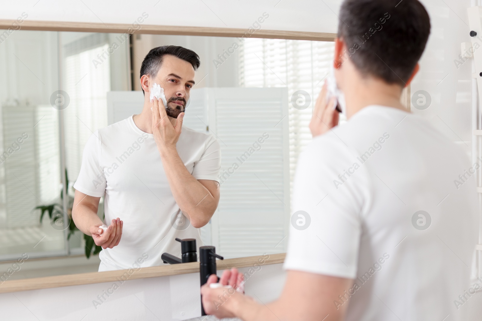 Handsome man applying shaving foam near mirror in bathroom Photo of Handsome man applying shaving foam near mirror in bathroom