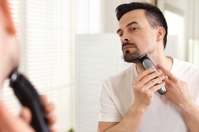 Photo of Handsome man shaving with trimmer near mirror at home