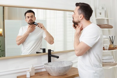 Photo of Handsome man shaving with trimmer near mirror at home