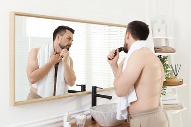 Photo of Handsome man shaving with trimmer near mirror in bathroom